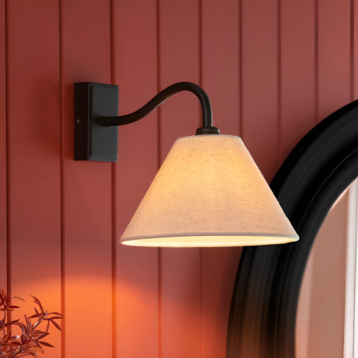 Endon Brockwood wall light with curved matt black arm and tapered natural linen shade, mounted on a red panelled wall beside a round black-framed mirror.