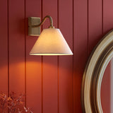 Brockwood Wall Light in brass with a curved arm and tapered natural linen shade, mounted on a red panelled wall beside a round mirror.