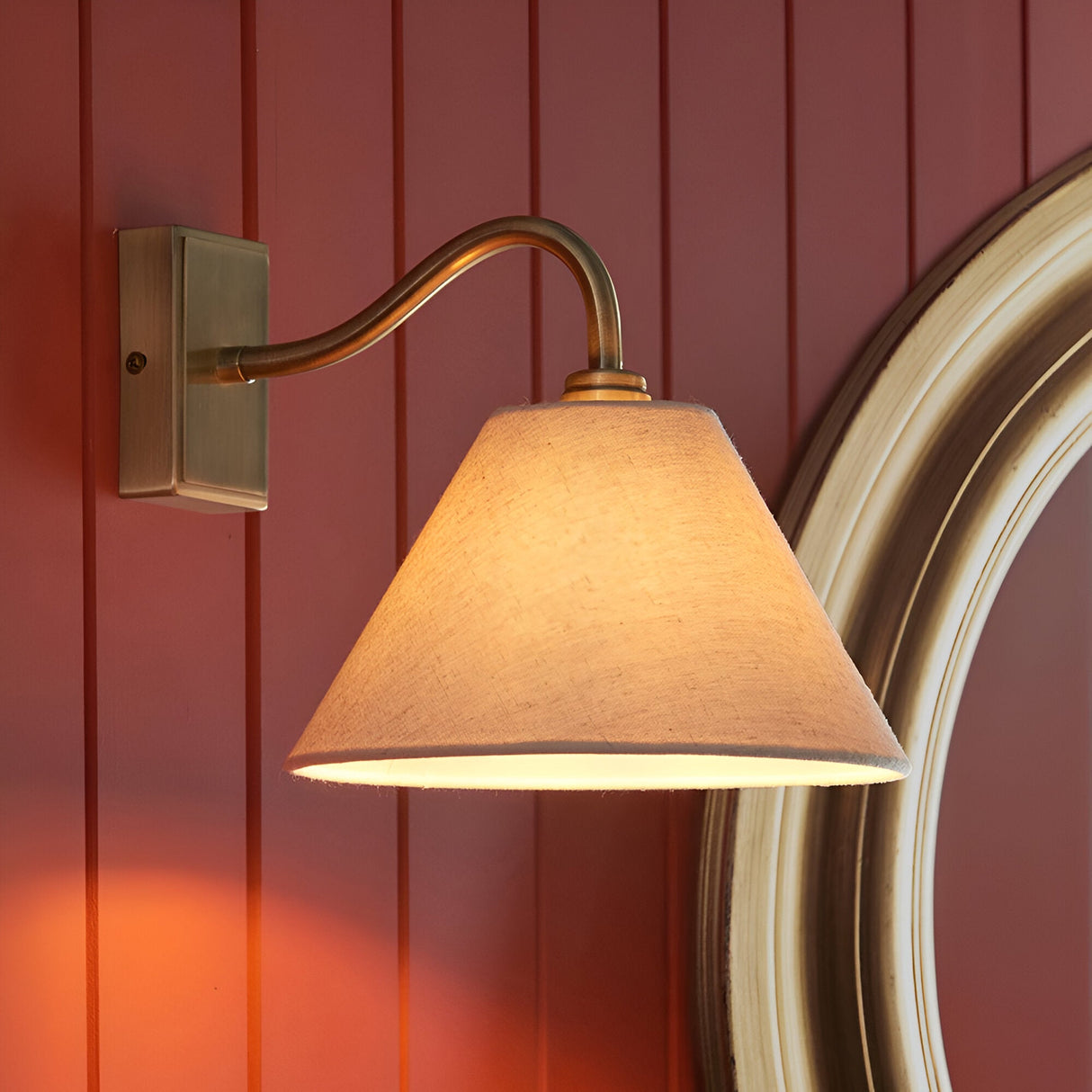 Brockwood Wall Light in brass with a curved arm and tapered natural fabric shade, mounted on a red panelled wall beside a mirror.