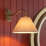 Brockwood Wall Light in brass with a curved arm and tapered natural fabric shade, mounted on a red panelled wall beside a mirror.