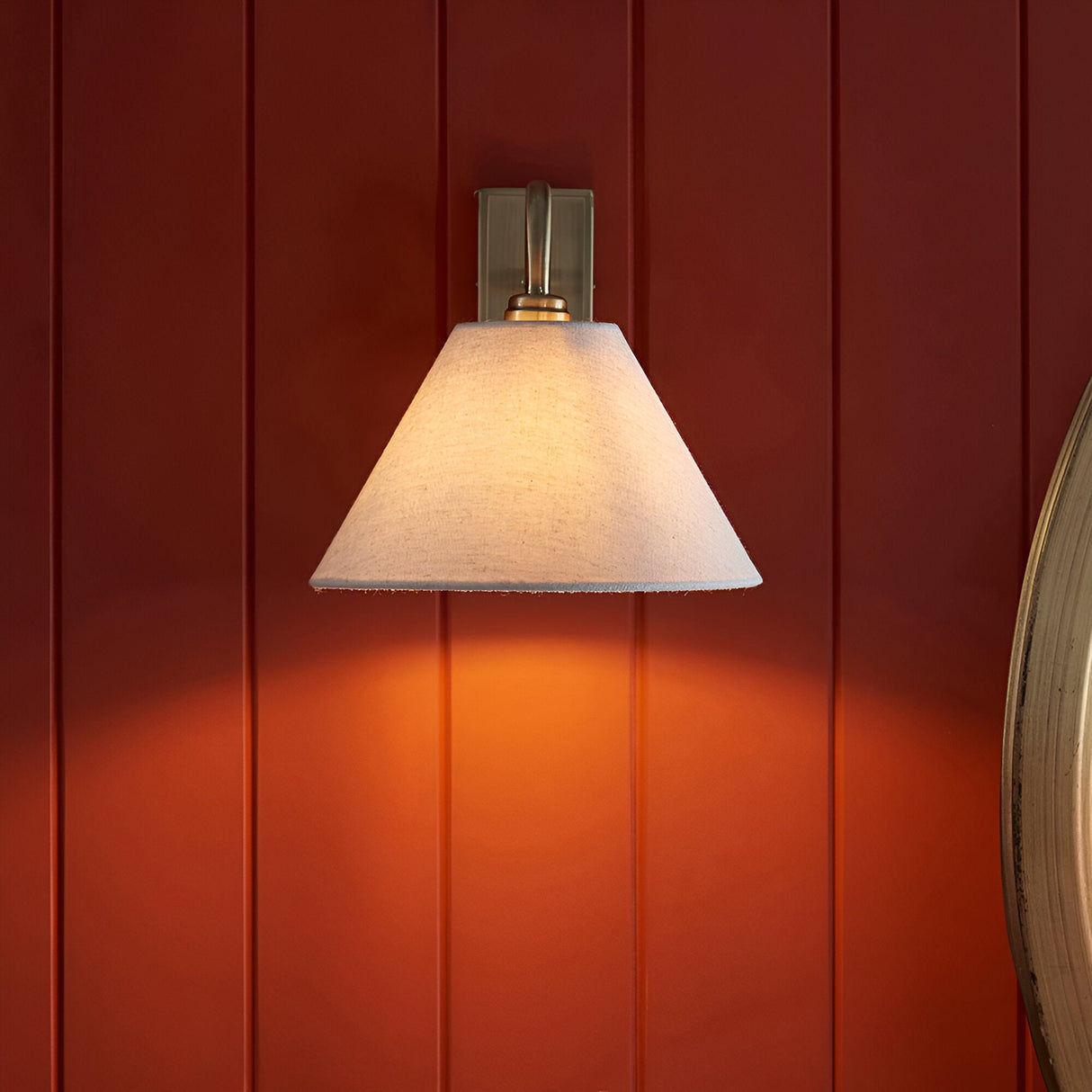 Brockwood Wall Light in brass with a curved metal arm and tapered natural linen shade, mounted on a dark red panelled wall for a warm ambient glow.