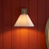 Brockwood Wall Light in brass with a curved metal arm and tapered natural linen shade, mounted on a dark red panelled wall for a warm ambient glow.