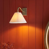 Brockwood Wall Light in brass with a tapered natural linen shade on a curved metal arm, mounted on a red panelled wall for a warm glow.