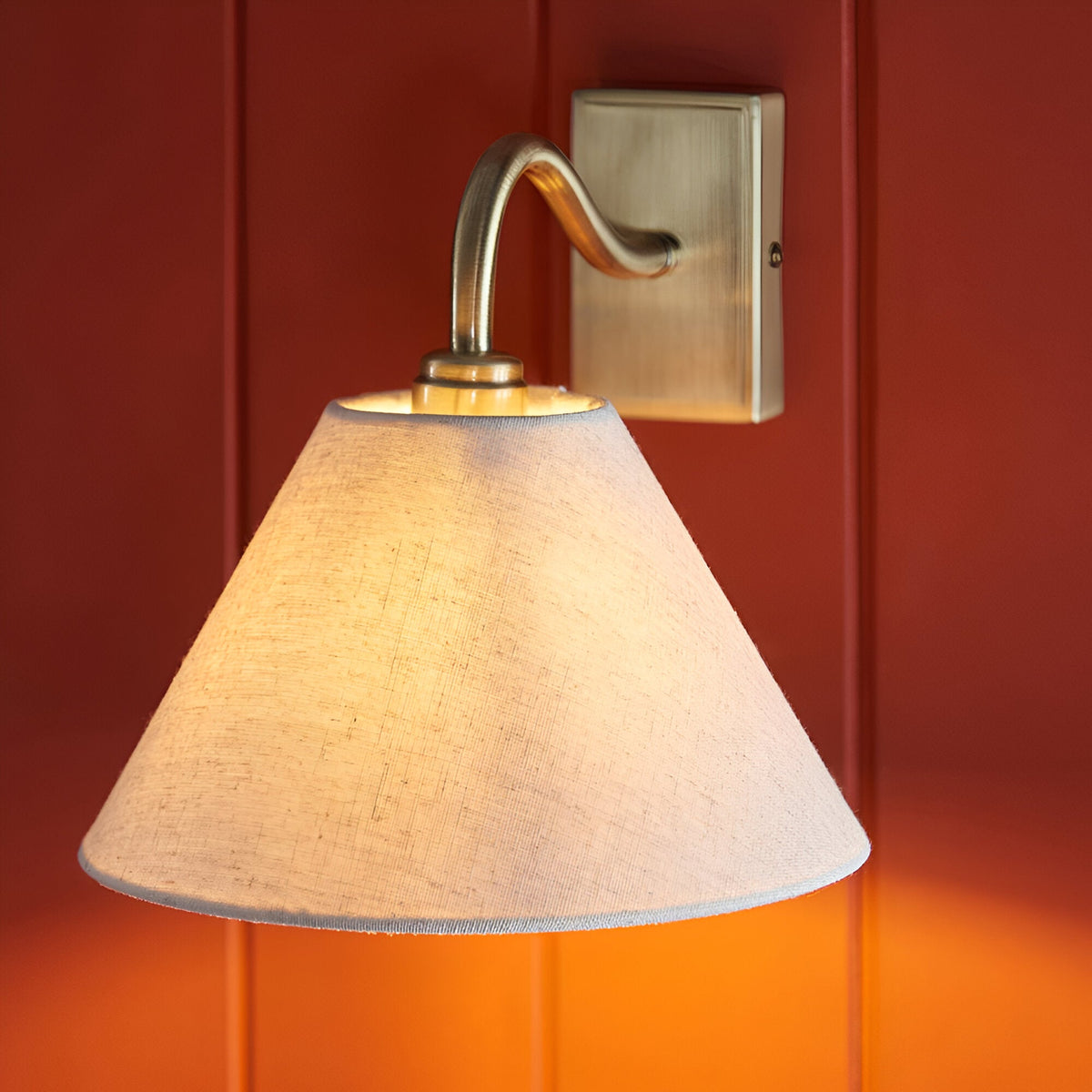 Brockwood Wall Light with curved brass arm and tapered natural linen shade, mounted on a red panelled wall with a warm glow.