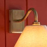 Brockwood Wall Light in brass with a curved metal arm and tapered natural linen shade, mounted on a rectangular brass backplate against a red panelled wall.