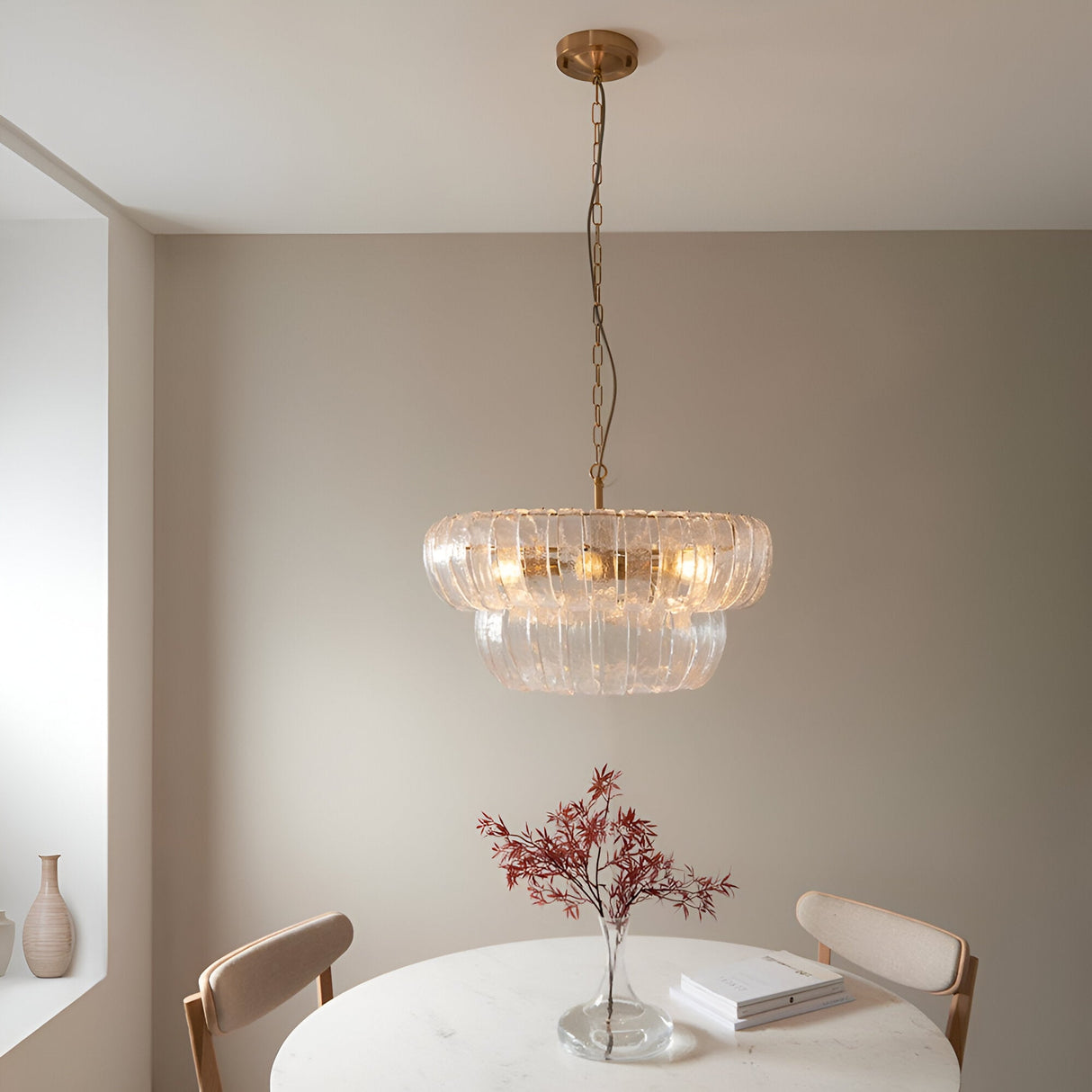 Carlwell 6 Light Pendant in brushed antique brass with two-tier curved clear glass, hung above a dining table in a neutral-toned room.