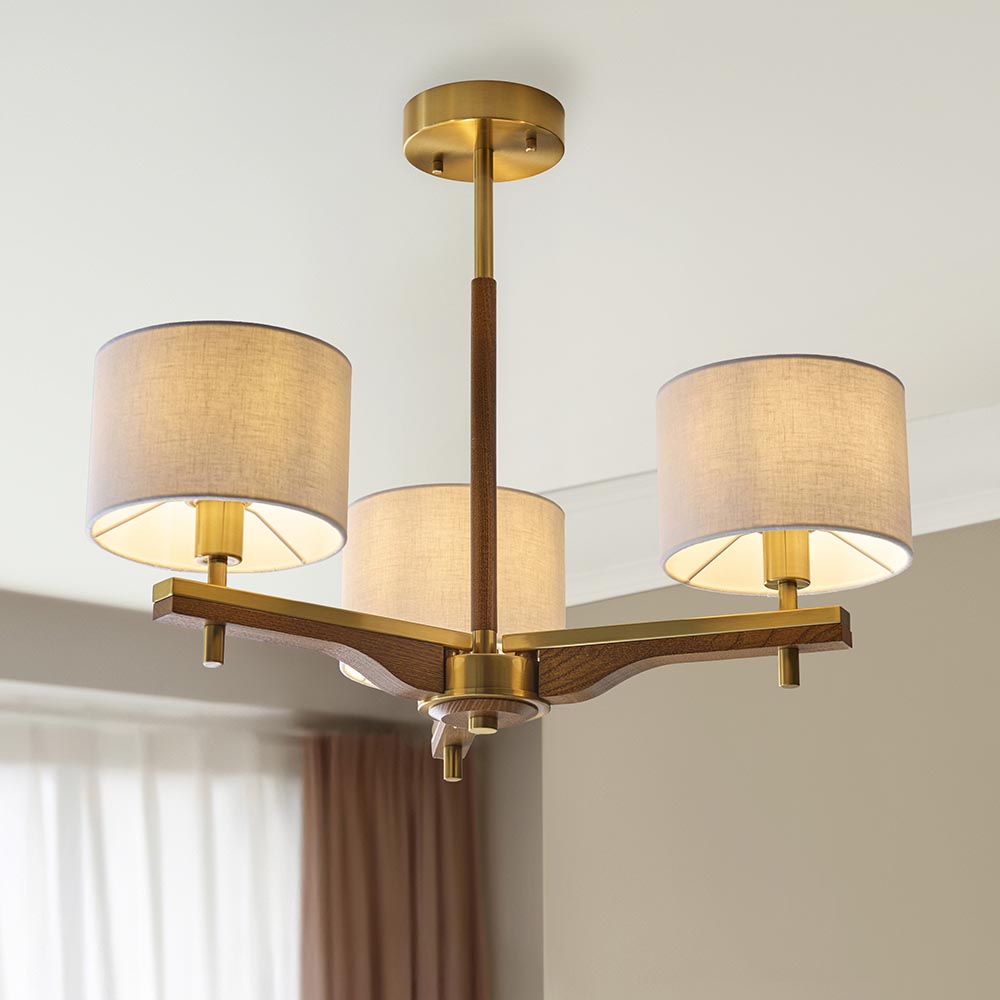 Bramwell 3-light semi-flush ceiling light with aged brass frame, oak arms and cream linen drum shades, shown fitted in a room with pale walls.