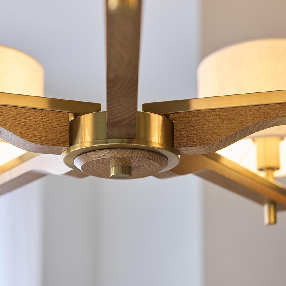 Close-up of the Bramwell 5 Light Pendant's centre hub, showing brushed brass fittings, oak-stained wood arms, and natural linen cylinder shades.