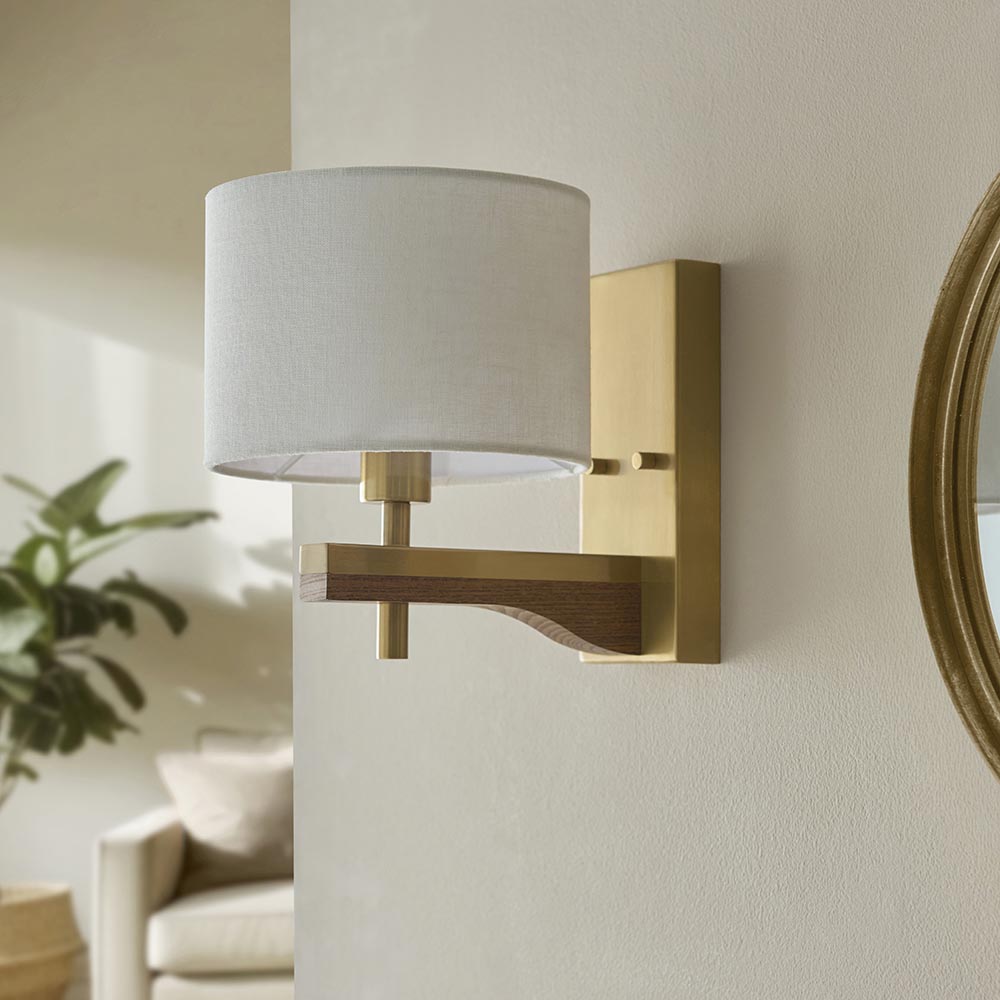 Endon Bramwell wall light in antique brushed brass with oak-stained wood arm and natural faux-linen cylinder shade, mounted on a pale wall beside a mirror.
