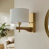Endon Bramwell wall light in antique brushed brass with oak-stained wood arm and natural faux-linen cylinder shade, mounted on a pale wall beside a mirror.