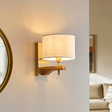 Endon Bramwell wall light in antique brushed brass with oak-stained wood bracket and natural faux-linen cylinder shade, lit on a wall beside a round mirror.