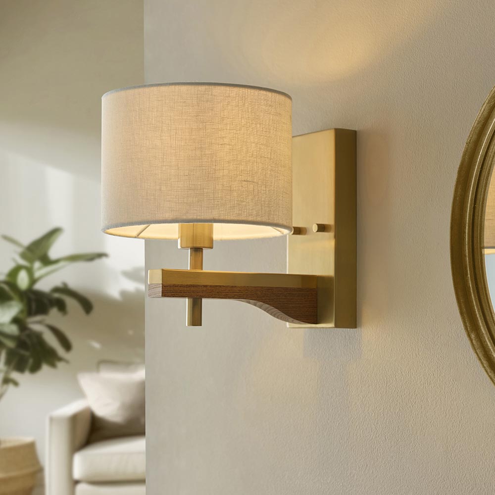 Endon Bramwell wall light in antique brushed brass with oak-stained wood arm and natural faux-linen cylinder shade, mounted beside a round gilt mirror.