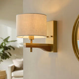 Endon Bramwell wall light in antique brushed brass with oak-stained wood arm and natural faux-linen cylinder shade, mounted beside a round gilt mirror.