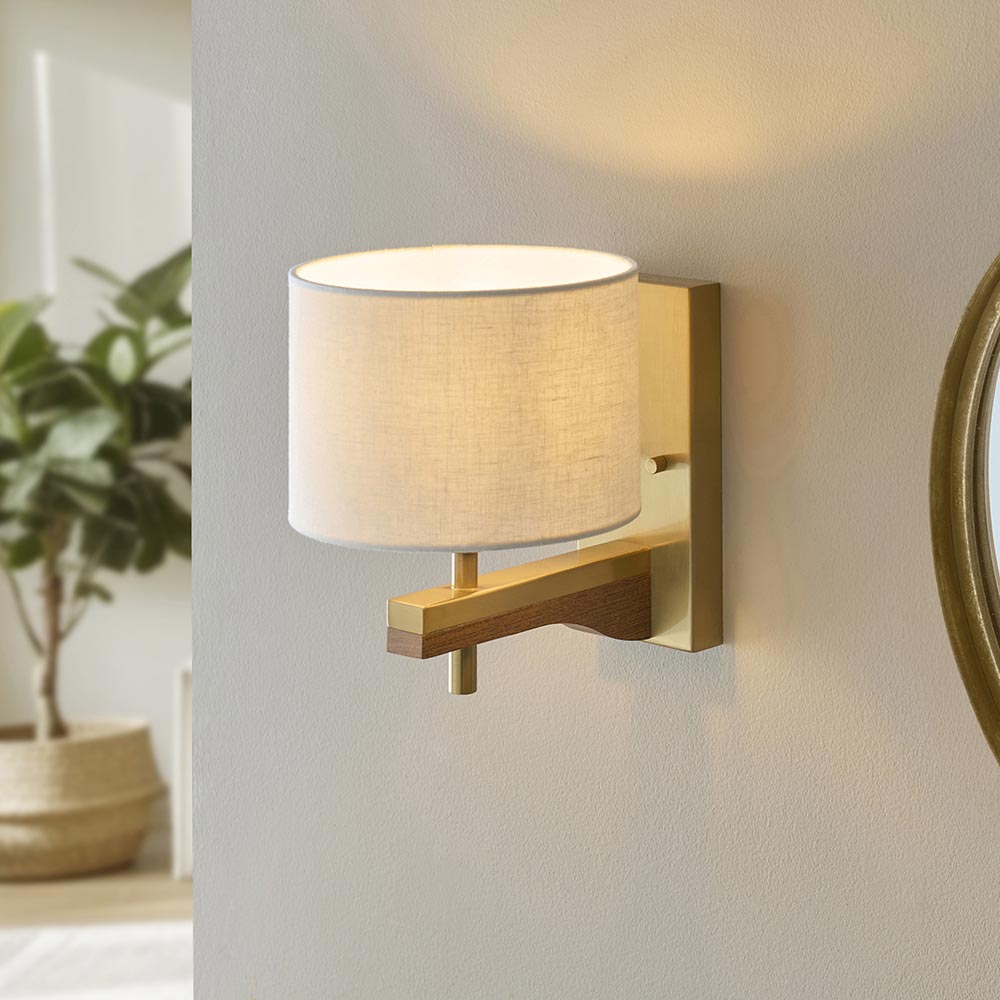 Endon Bramwell wall light with oak-stained ash arm, brushed antique brass mount and natural faux-linen cylinder shade, lit on a grey wall beside a mirror.