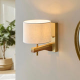 Endon Bramwell wall light with oak-stained ash arm, brushed antique brass mount and natural faux-linen cylinder shade, lit on a grey wall beside a mirror.