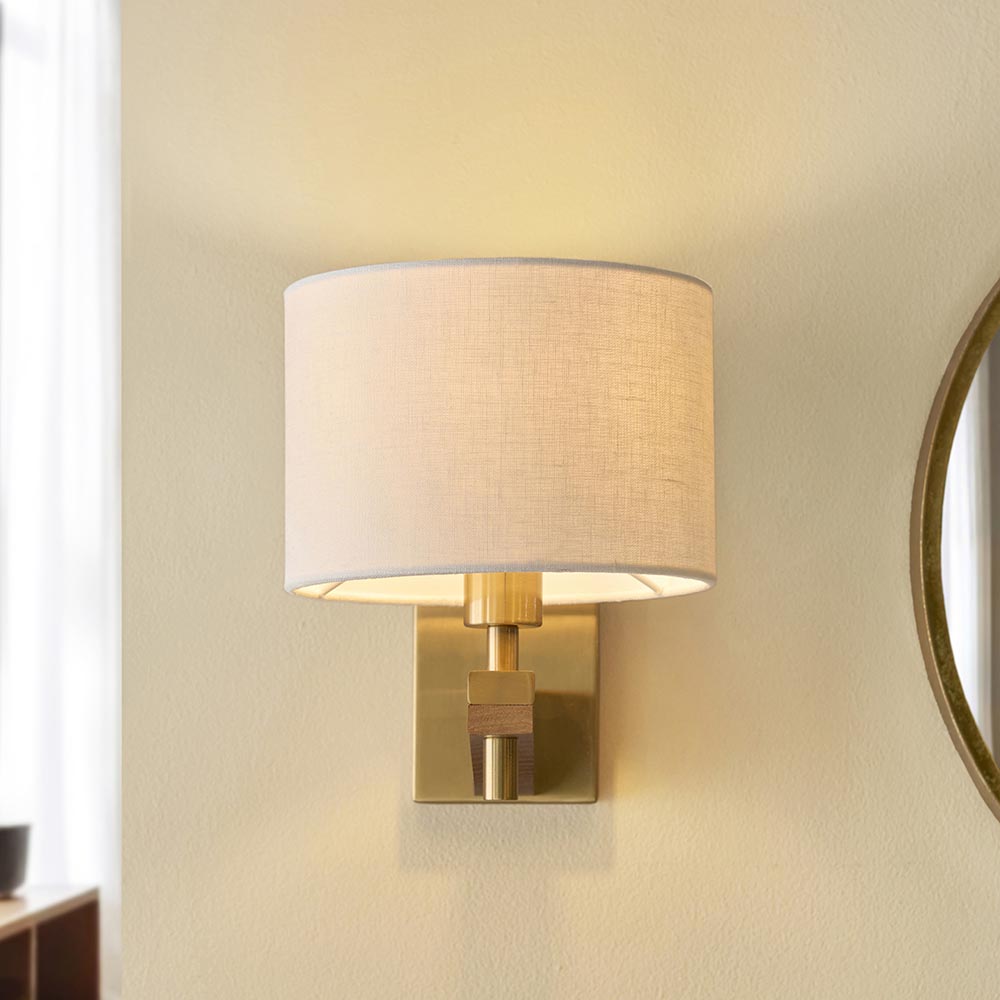 Endon Bramwell wall light in antique brushed brass with oak-stained wood and faux-linen cylinder shade, mounted in a hallway beside a round mirror.