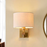 Endon Bramwell wall light in antique brushed brass with oak-stained wood and faux-linen cylinder shade, mounted in a hallway beside a round mirror.
