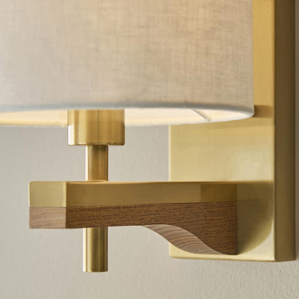 Close-up of the Endon Bramwell wall light showing brushed antique brass metalwork, oak-stained ash wood detail, and the base of a natural faux-linen shade.