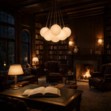 Cozy library with leather chairs, bookshelves, a lit fireplace, and the Auriel 7 Light Alabaster Chandelier in Soft White & Brushed Brass glowing overhead. An open book lies on a large wooden table in the foreground.