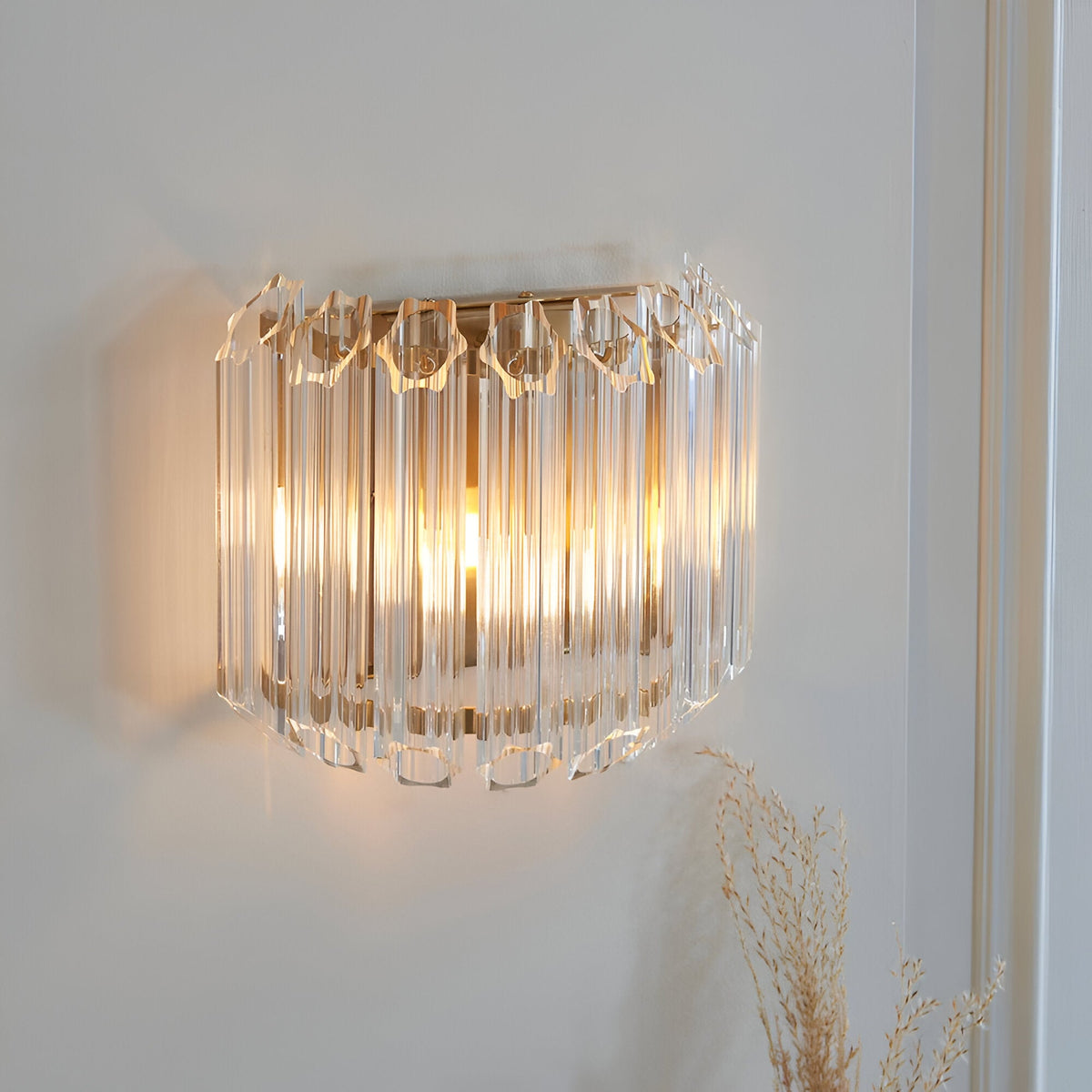 Endon Danebury wall light with faceted clear glass rods on a brushed aged brass frame, illuminated on a pale wall beside dried grasses.