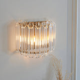 Endon Danebury wall light with faceted clear glass rods on a brushed aged brass frame, illuminated on a pale wall beside dried grasses.