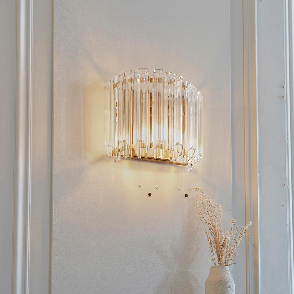 Endon Danebury wall light in brushed aged brass with faceted clear glass rods, lit on a white panelled wall above a vase of dried grasses.