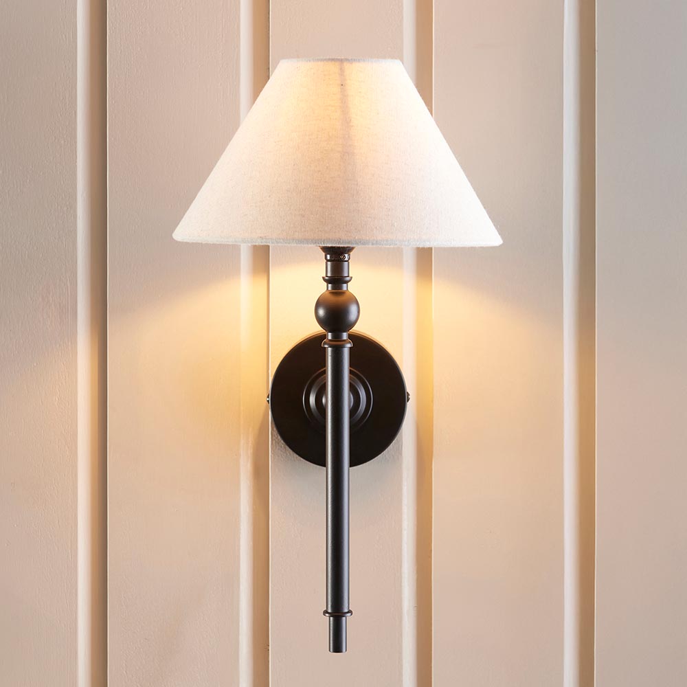 Endon Caldmere wall light in matt black with tapered natural linen shade, mounted on a cream panelled wall and casting a warm glow.