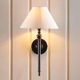 Endon Caldmere wall light in matt black with tapered natural linen shade, mounted on a cream panelled wall and casting a warm glow.