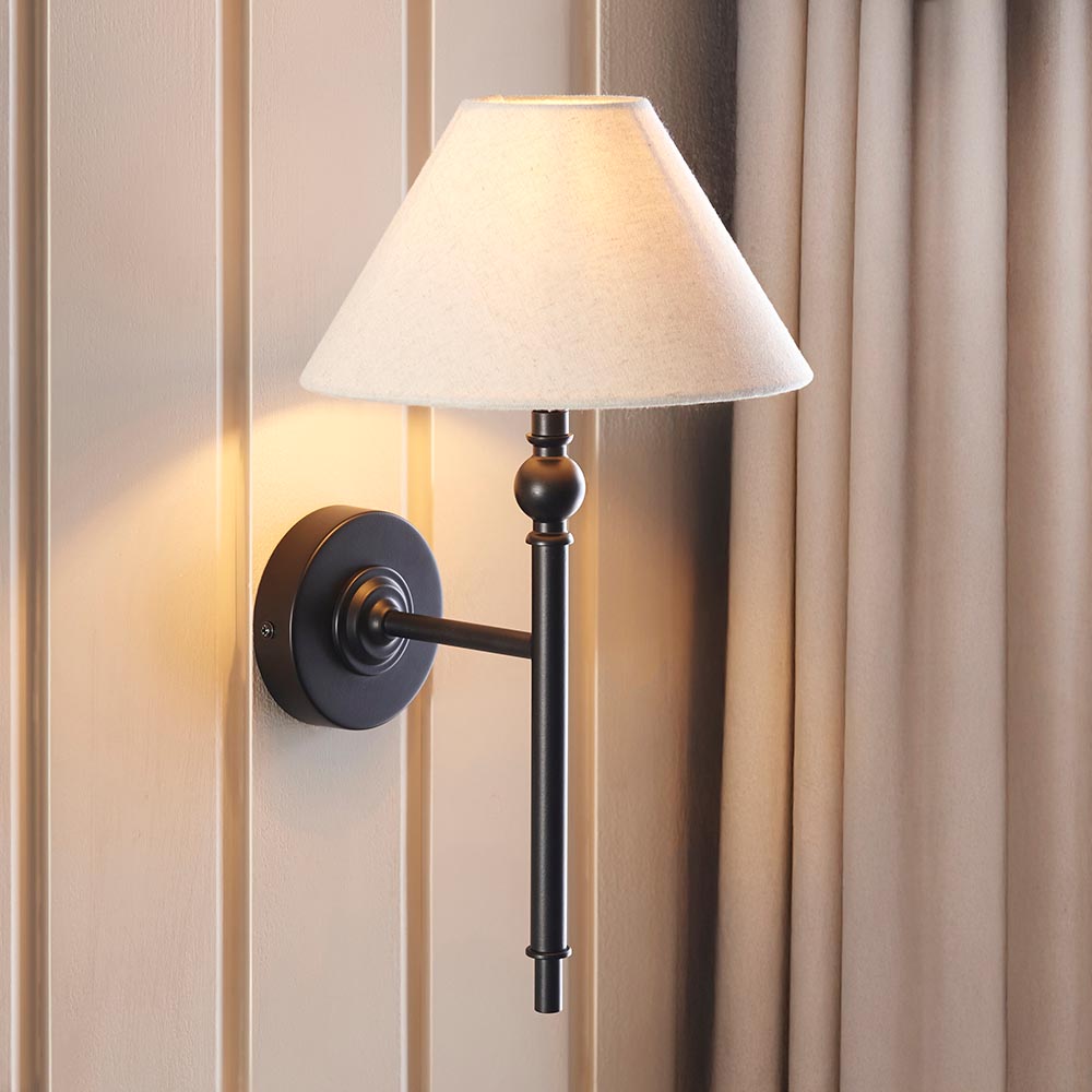 Endon Caldmere wall light in matt black with tapered natural linen shade, mounted on a panelled wall beside soft beige curtains.