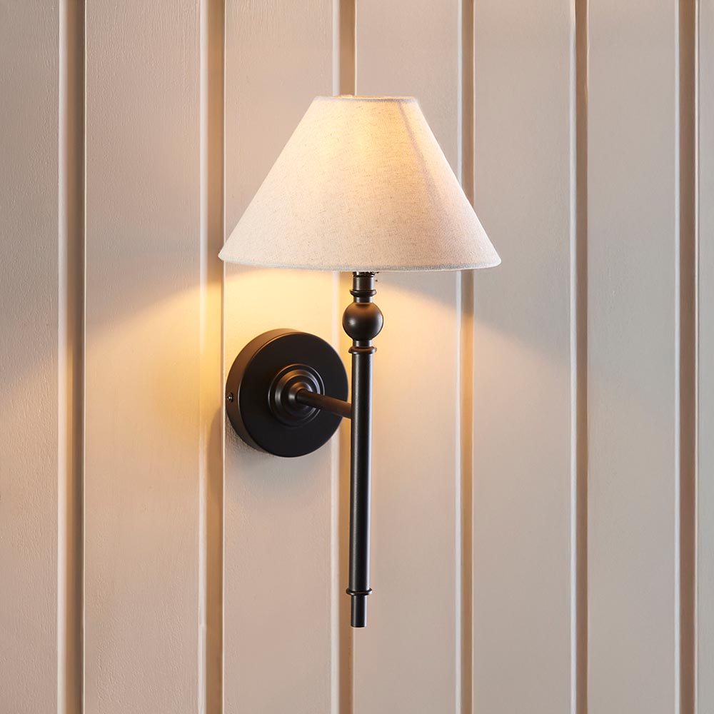 Endon Caldmere wall light in black with a tapered natural linen shade, mounted on a cream panelled wall casting a warm glow.