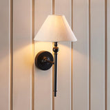 Endon Caldmere wall light in black with a tapered natural linen shade, mounted on a cream panelled wall casting a warm glow.