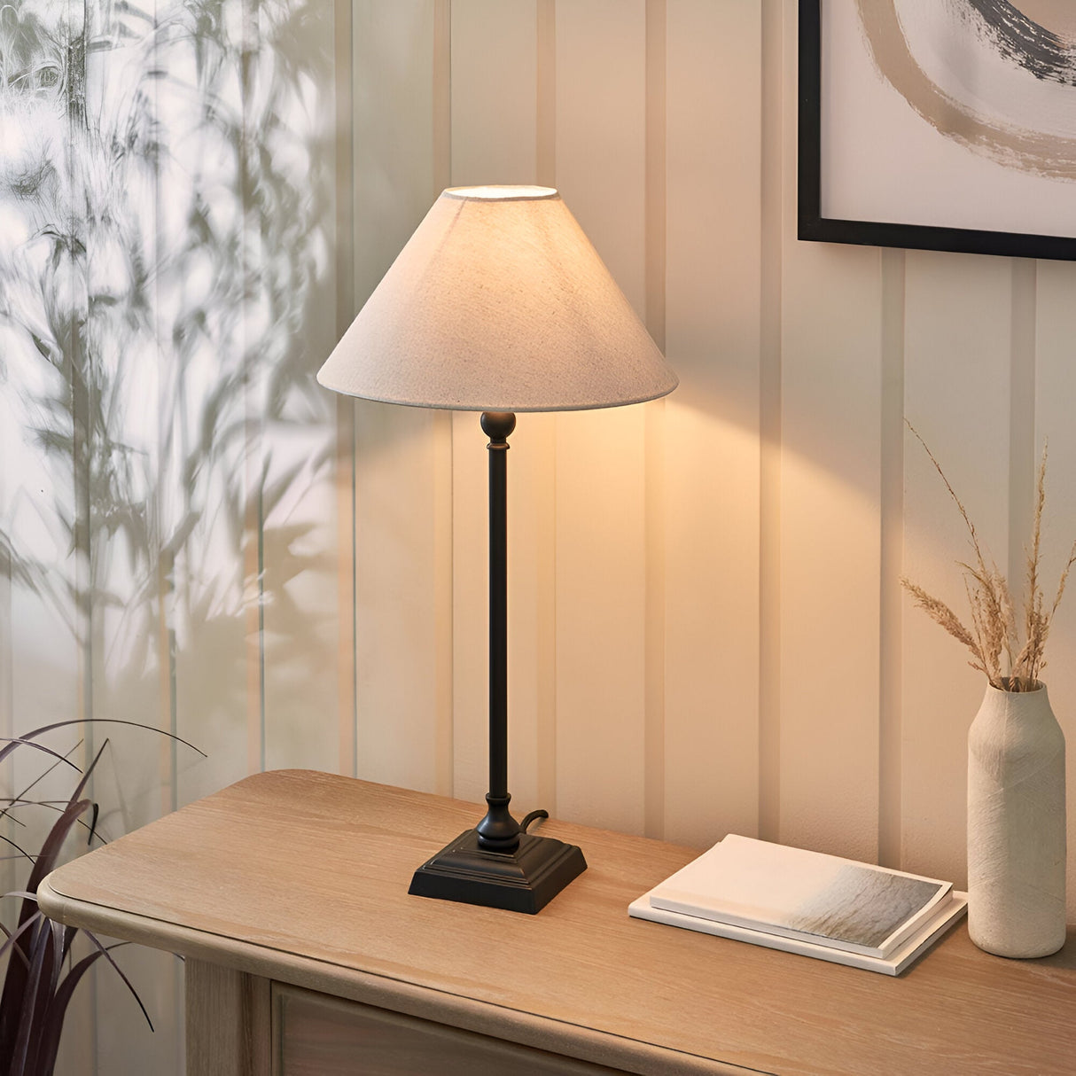 Endon Caldmont table lamp with slim matt black turned metal column and natural linen tapered shade, lit on a wooden console beside a book and vase.