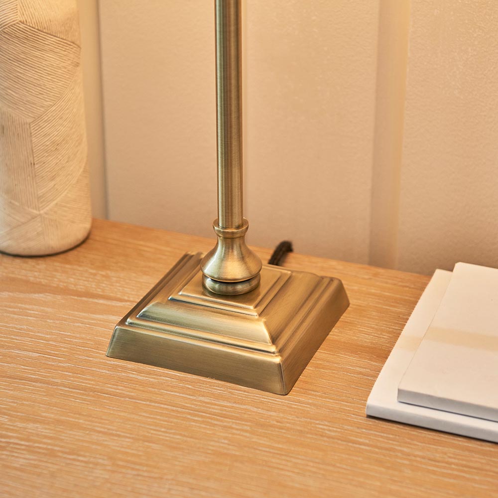 Caldmont Table Lamp base close-up in brass, with stepped square foot and slim turned column, shown on a wooden bedside table.