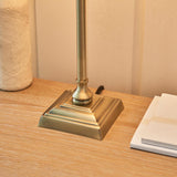 Caldmont Table Lamp base close-up in brass, with stepped square foot and slim turned column, shown on a wooden bedside table.