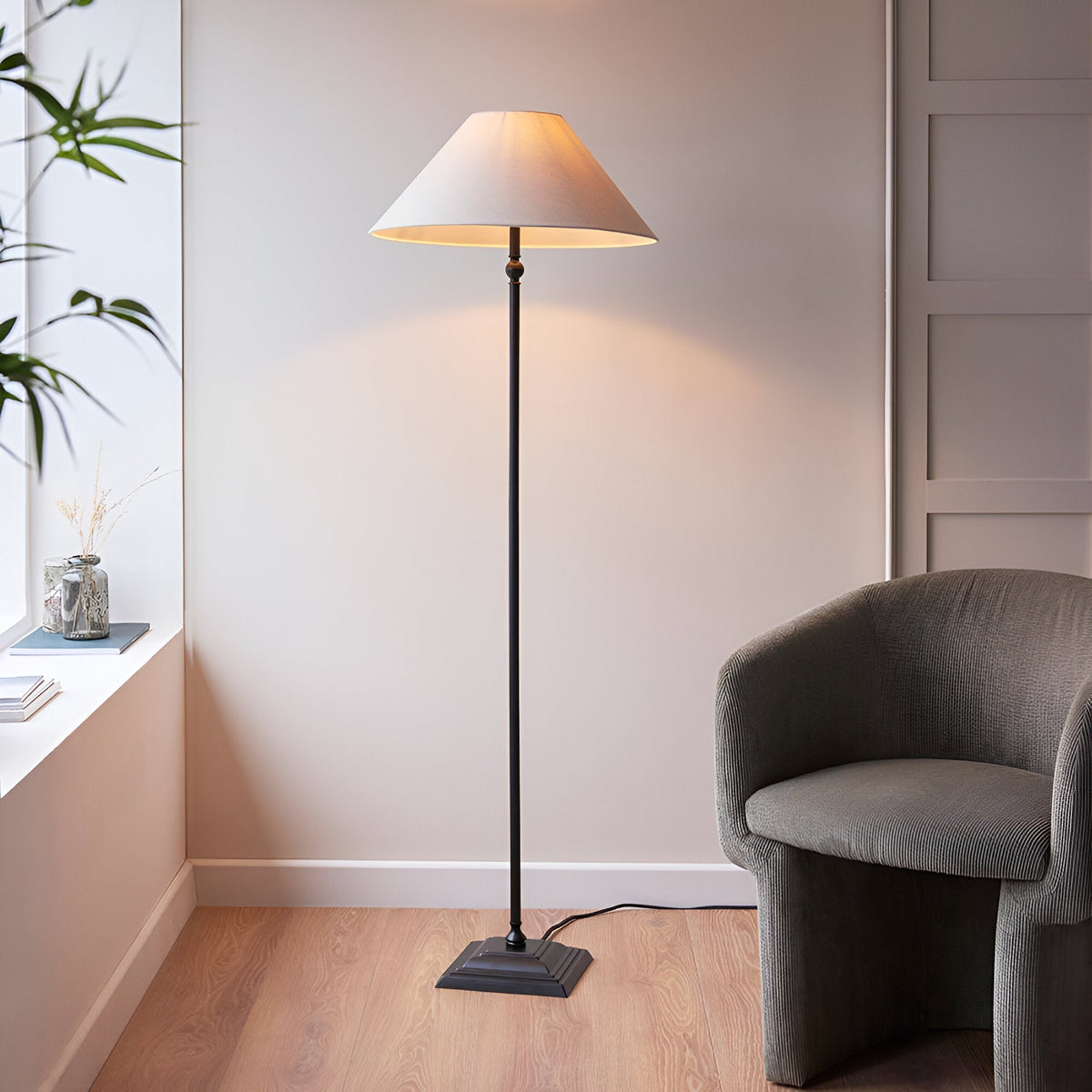 Endon Caldmere floor lamp with tapered natural linen shade and matt black metal stem on a stepped base, lit beside a grey armchair in a living room.