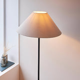 Endon Caldmere floor lamp with tapered natural linen shade and matt black metal stem, lit beside a windowsill with a glass vase of dried grasses.