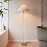 Endon Caldmere floor lamp in antique brass with a tapered natural linen shade, lit beside a grey armchair in a neutral living room.
