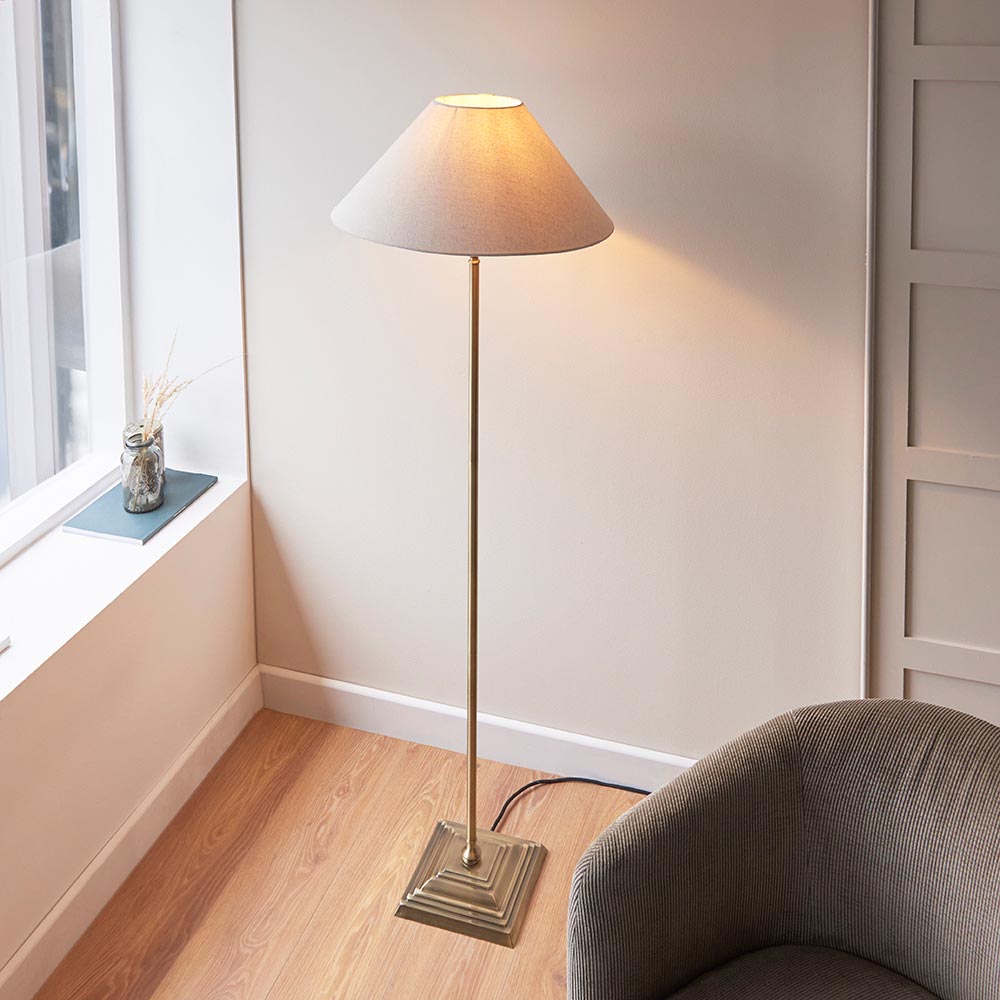 Endon Caldmere floor lamp in antique brass with a tapered natural linen shade, beside a grey armchair and window in a living room corner.