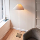 Endon Caldmere floor lamp in antique brass with a tapered natural linen shade, beside a grey armchair and window in a living room corner.
