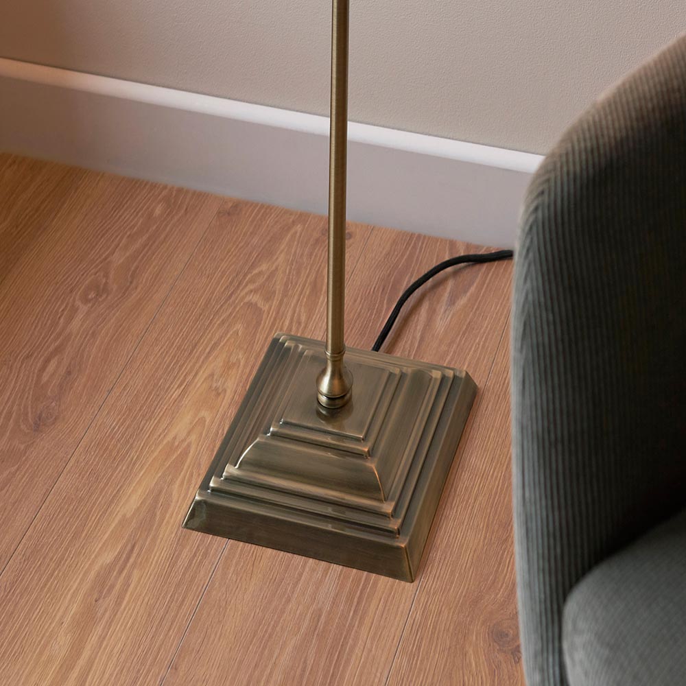 Endon Caldmere floor lamp base in antique brass with tiered stepped square plinth and slim stem, set on wood flooring beside a grey corduroy armchair.