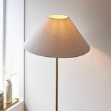 Endon Caldmere floor lamp in antique brass with a wide tapered natural linen shade, illuminated beside a windowsill with a dried grass vase.