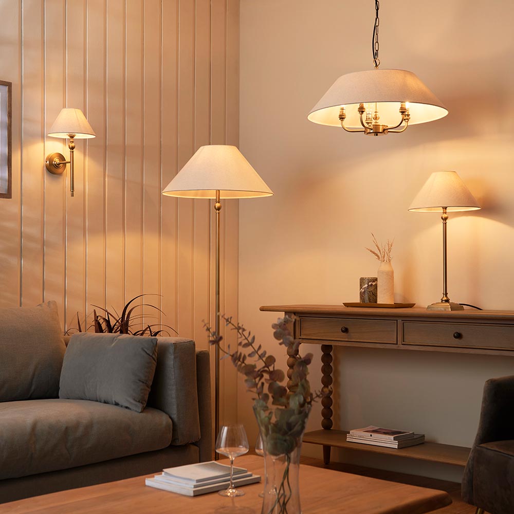 Endon Caldmere floor lamp with a tapered natural linen shade on a slim brass stem, shown beside a sofa in a warmly lit living room with matching lamps.