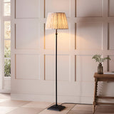 Caldmere Ikat Floor Lamp in Natural, featuring a matt black metal stem and base with a pleated Ikat fabric shade, shown lit beside a wood-panelled wall.