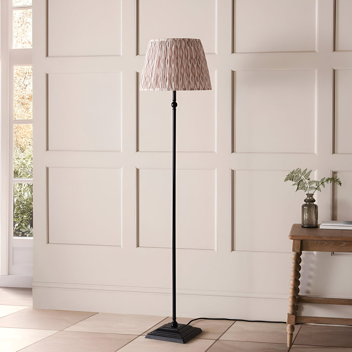 Caldmere Ikat Floor Lamp in Natural, featuring a pleated brown printed fabric shade on a matt black stepped metal base, shown in a panelled room.