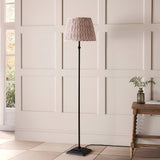 Caldmere Ikat Floor Lamp in Natural, featuring a pleated brown printed fabric shade on a matt black stepped metal base, shown in a panelled room.
