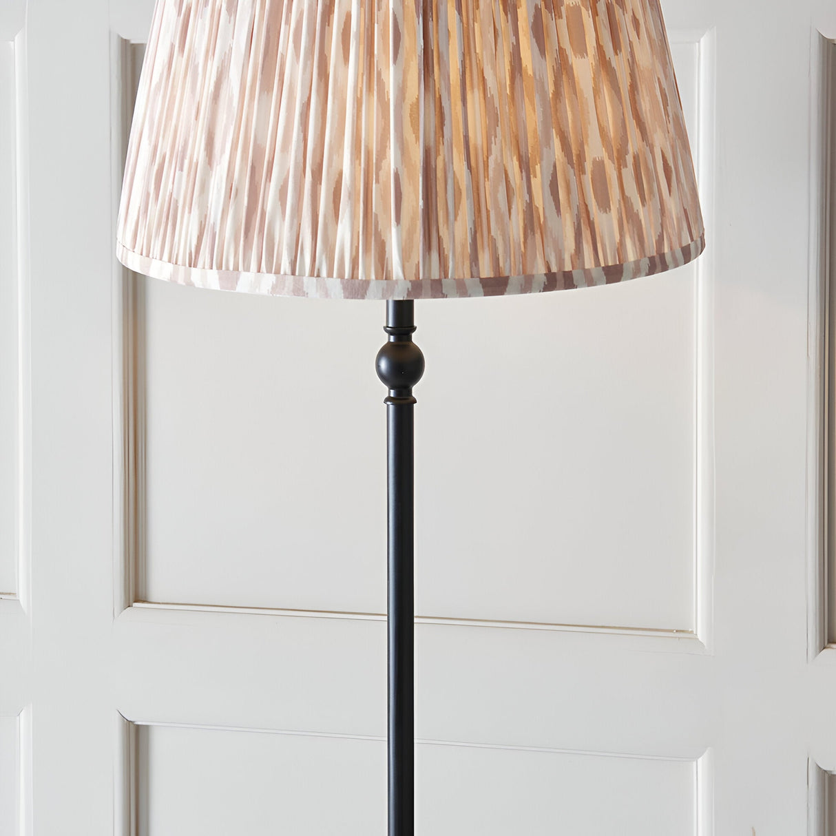 Caldmere Ikat Floor Lamp in natural fabric with a pleated 40cm shade on a matt black metal stem base, shown against a panelled white wall.