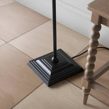 Caldmere Ikat Floor Lamp base close-up, showing matt black stepped square base and pole, on tiled floor beside a wooden table leg.