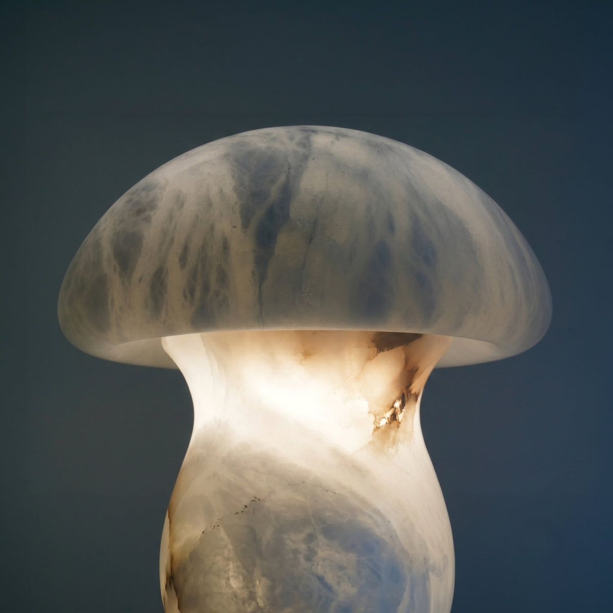 Morfia mushroom-form alabaster table lamp with domed cap, glowing warm white against a dark blue background, showing natural grey-brown veining.