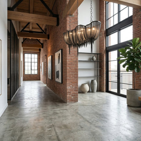 A modern loft with exposed brick, large windows, and wood beams. Polished concrete floors reflect natural light, while artworks adorn the walls. The Mireya 8 Light Metal Chain Veil Suspension Light - Black serves as a striking chandelier above.