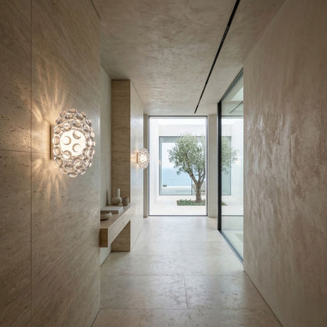A minimalist hallway features beige stone walls and floors, the Briller LED Small Wall Light - Transparent, a small bench with decor, and a large window showcasing a potted tree and distant ocean views.